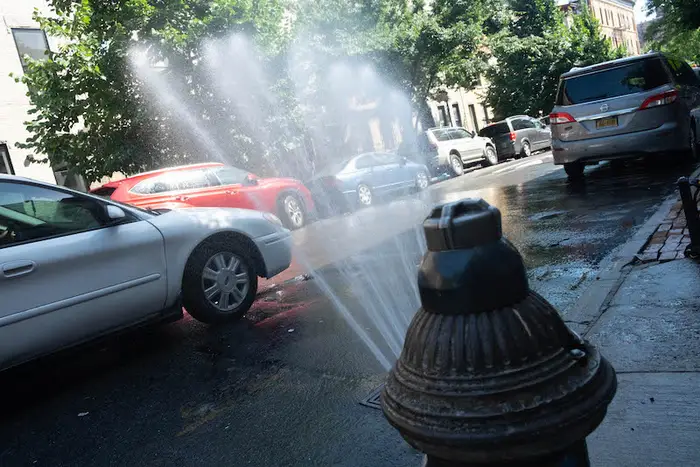 A silver sedan drives past an open fire hydrant on a Brooklyn street.